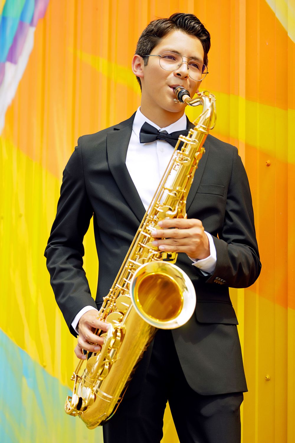 Musician in tuxedo plays saxophone against vibrant orange and yellow background.