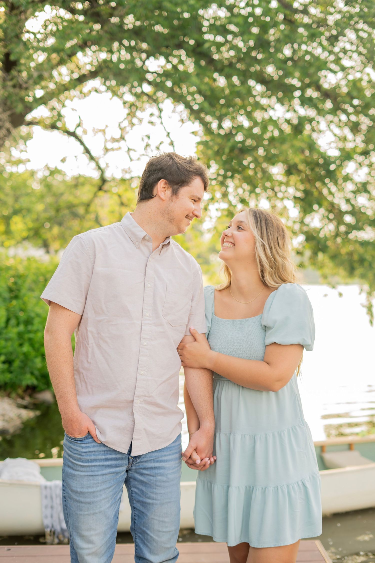 Engaged couple laughing and holding hands on a wooden dock with a river backdrop.