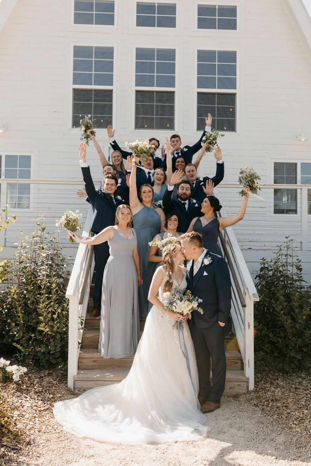 Bride and groom share a kiss on steps as joyful wedding party celebrates with raised bouquets behind them.