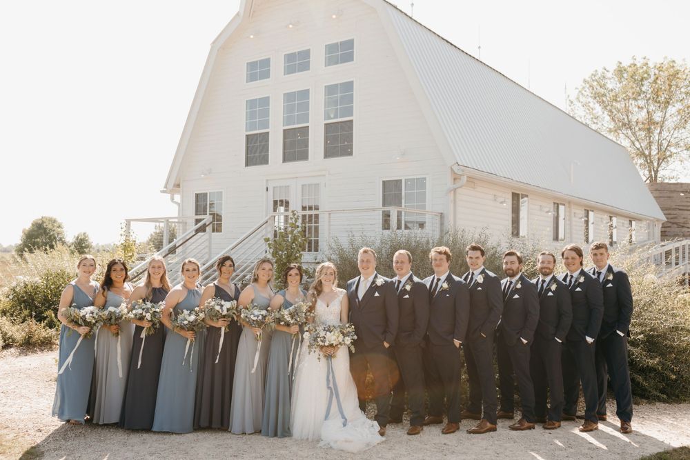 Large wedding party poses in front of a white barn venue with bridesmaids in dusty blue gowns.