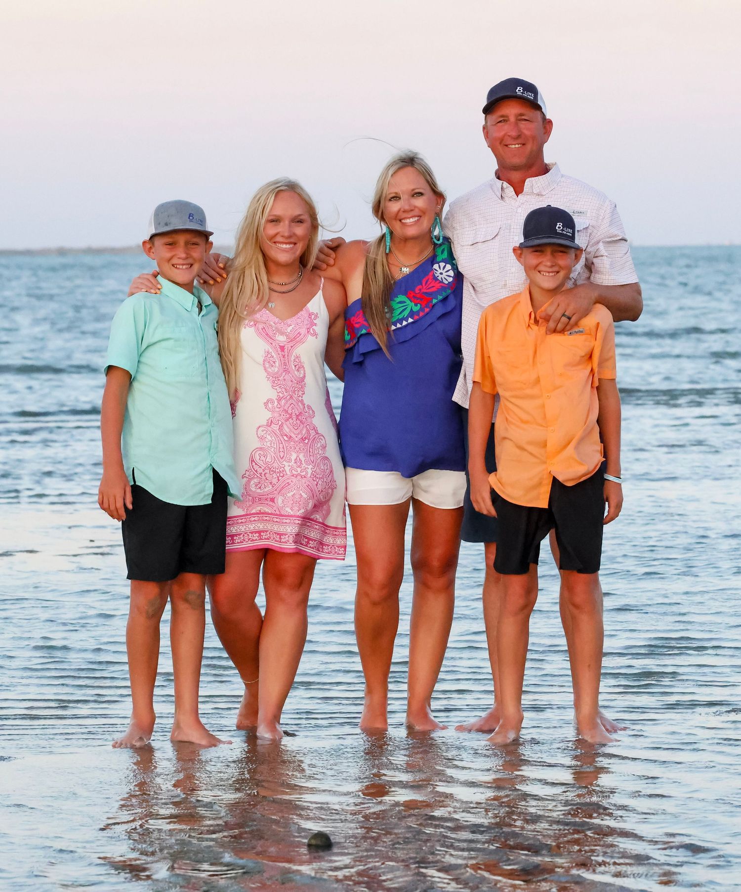 Group of people standing in shallow ocean water at sunset wearing colorful beach attire.