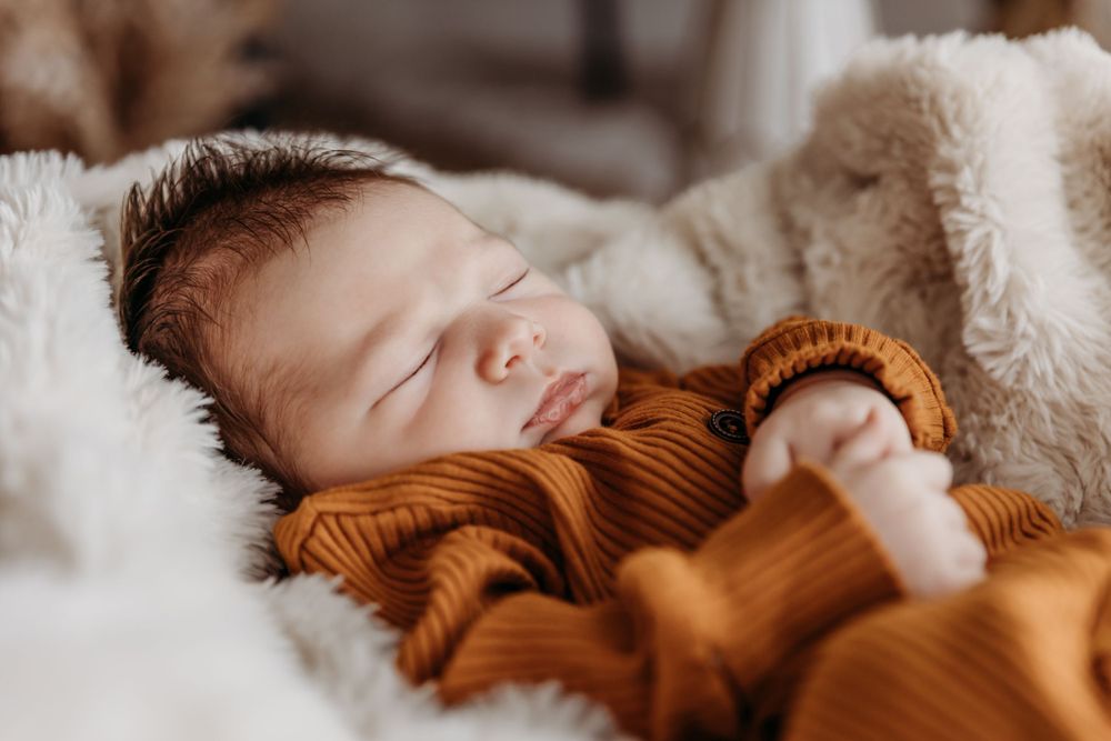 Newborn baby sleeping peacefully in cozy rust-colored outfit on white fur.