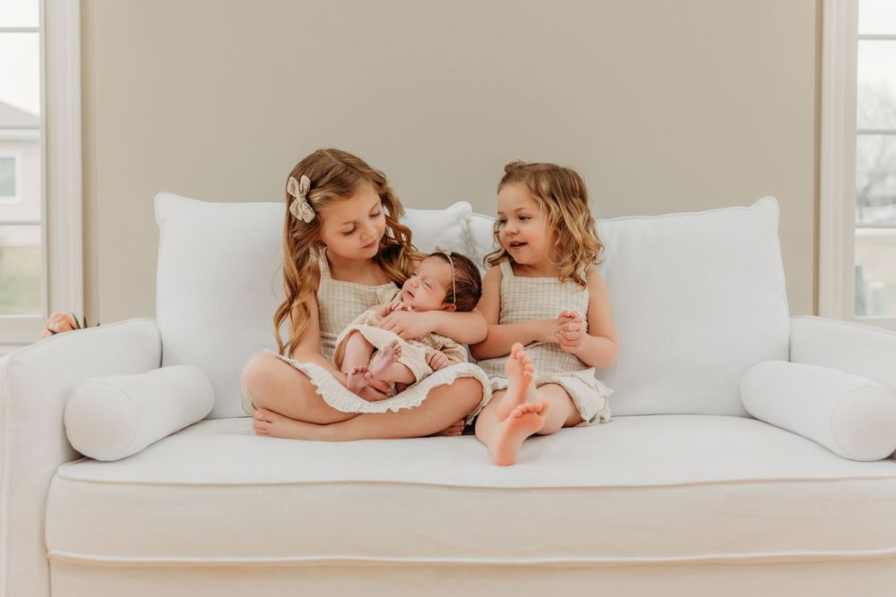 Three children sit together on a white couch sharing a tender moment with a newborn.