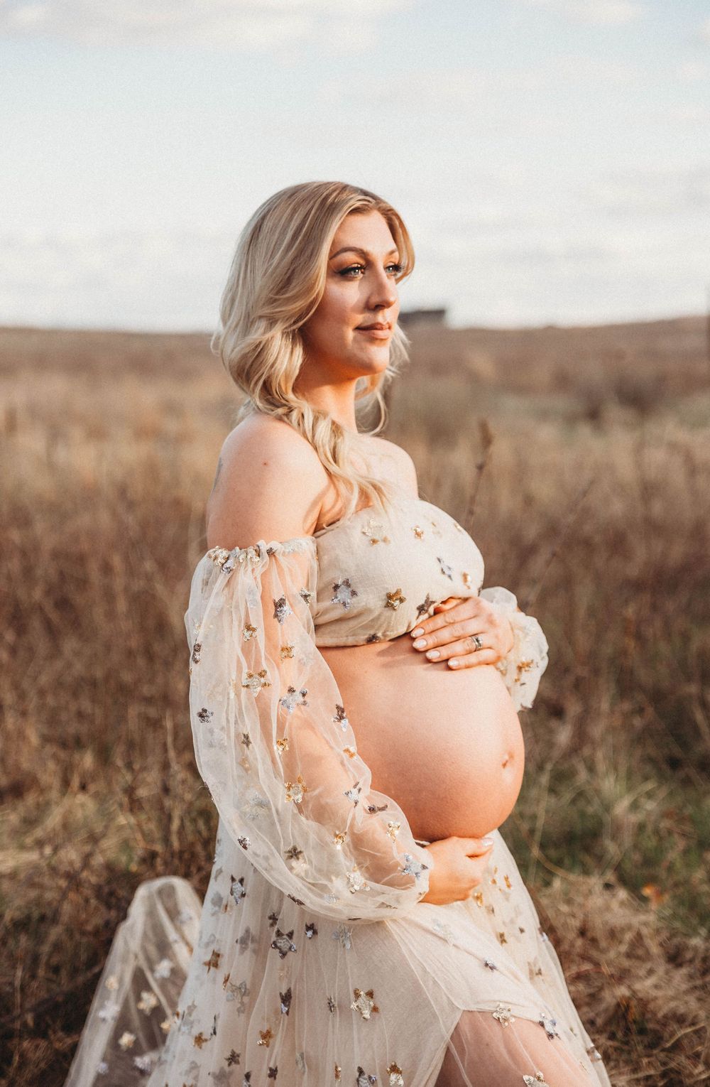 Maternity portrait in a delicate white lace dress against a natural outdoor setting with warm lighting.