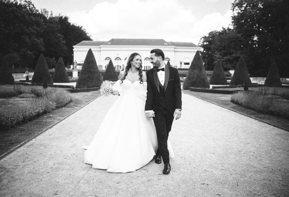 Black and white wedding photo of couple walking along elegant garden path with classic architecture.