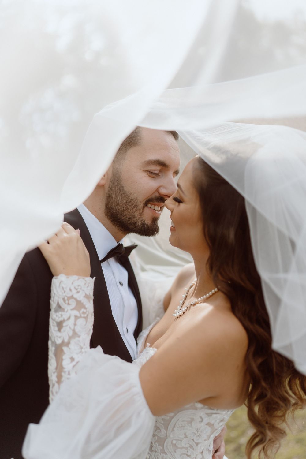Romantic wedding portrait under white veil with delicate lace details and pearl necklace.