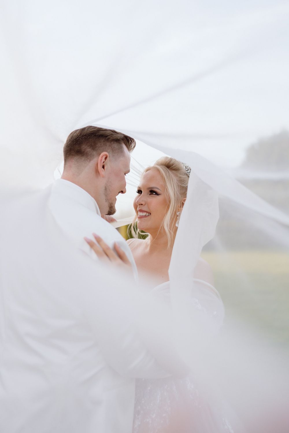 Romantic wedding portrait series of a couple embracing under flowing white veil in soft natural light.