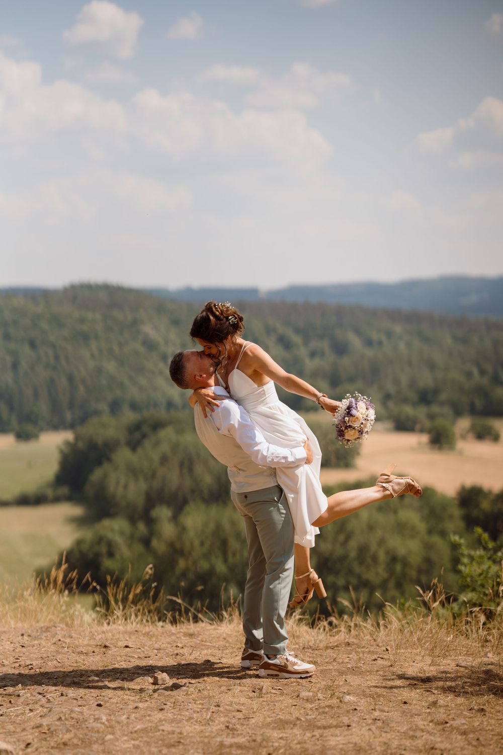 Playful wedding couple shares joyful moment on hilltop with scenic valley view.