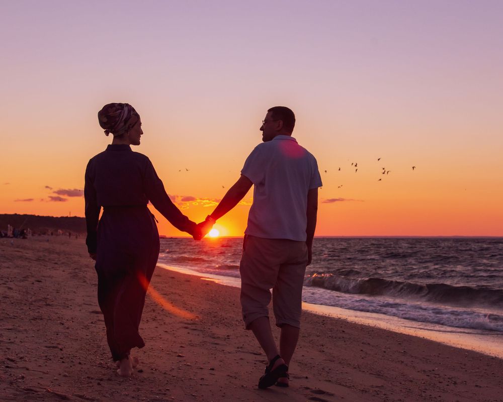 Silhouetted couple walking hand in hand on a beach at sunset with birds flying in the orange sky.