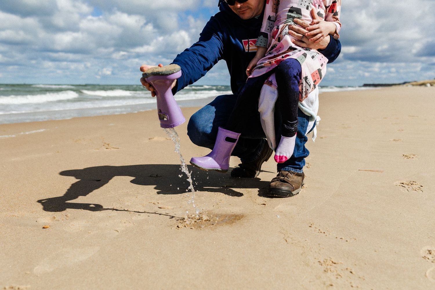 a dad holding his daughter and dumping water out of her boots on the beach