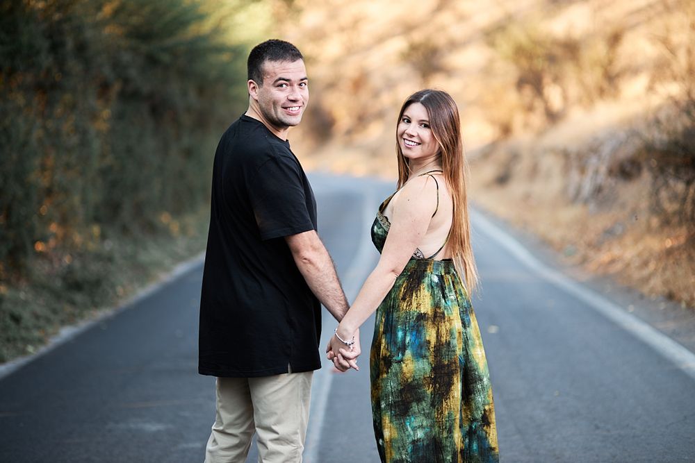 A couple looks back at camera while holding hands on a rural road at sunset.