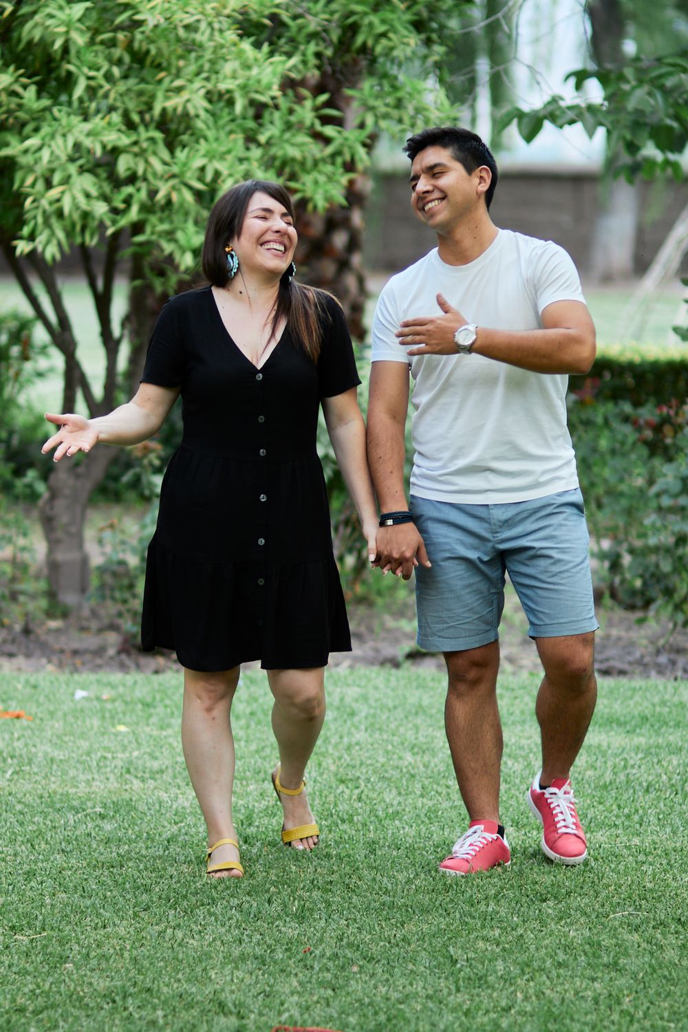A couple laughs together while walking on grass with trees in the background.