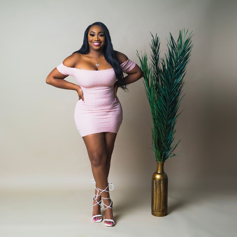 Studio portrait of a curvy model in a pink mini dress posing next to a tall tropical plant in a gold vase.