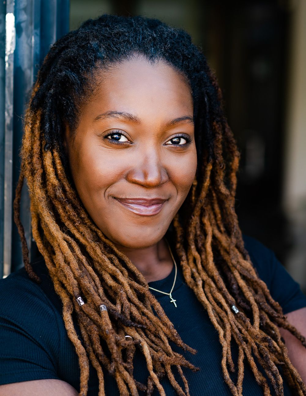 Portrait of a person with warm-toned locs wearing a black t-shirt against a dark background.