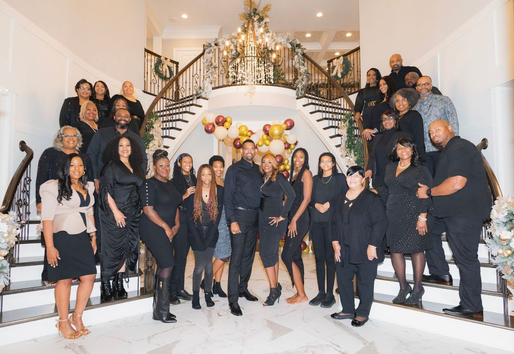 Large group photo of professionals in black attire posing on an elegant white staircase with chandelier.