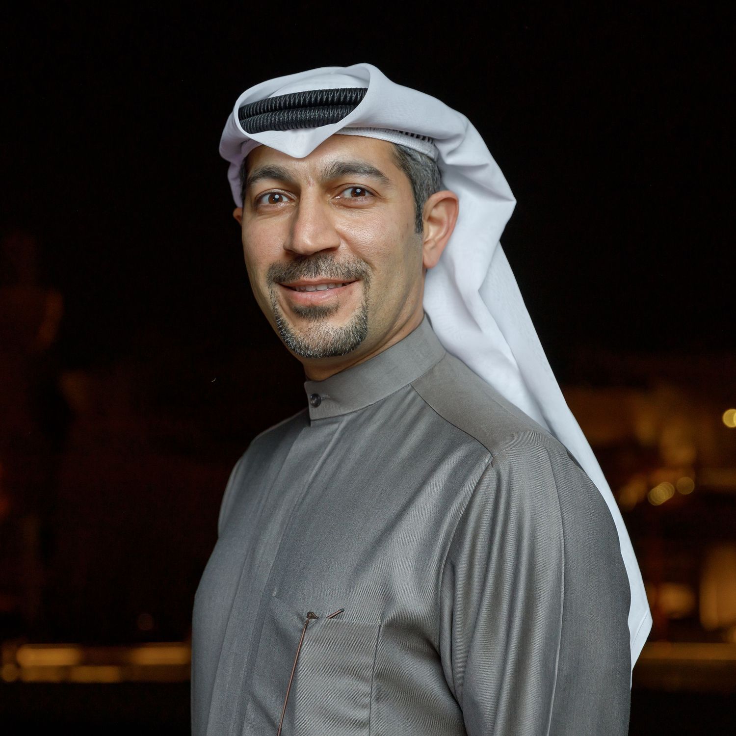 Man in traditional Gulf dress and headwear poses against dark background with lit buildings.