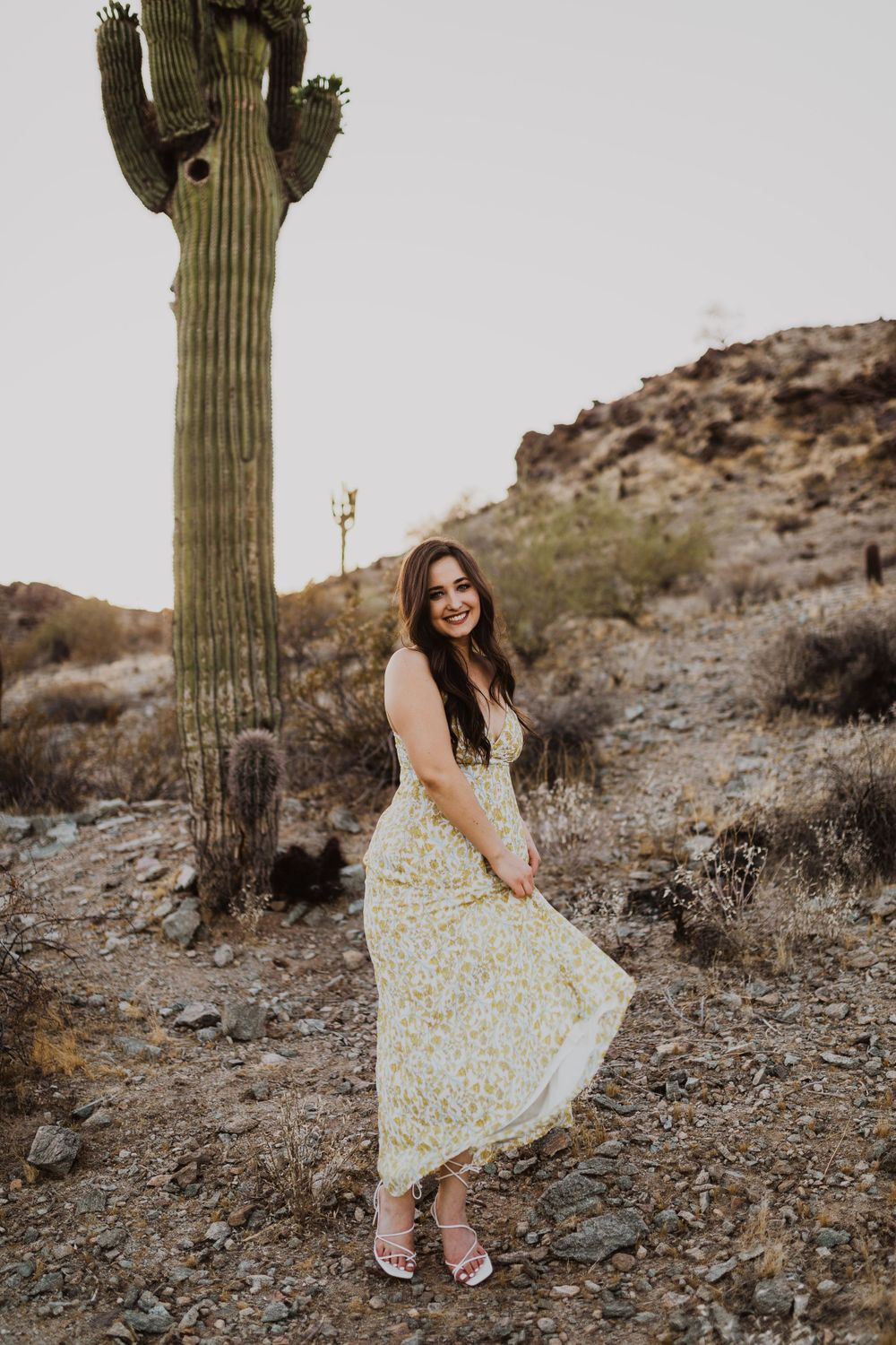 Fashion model in floral dress poses playfully near giant saguaro cactus in desert setting.