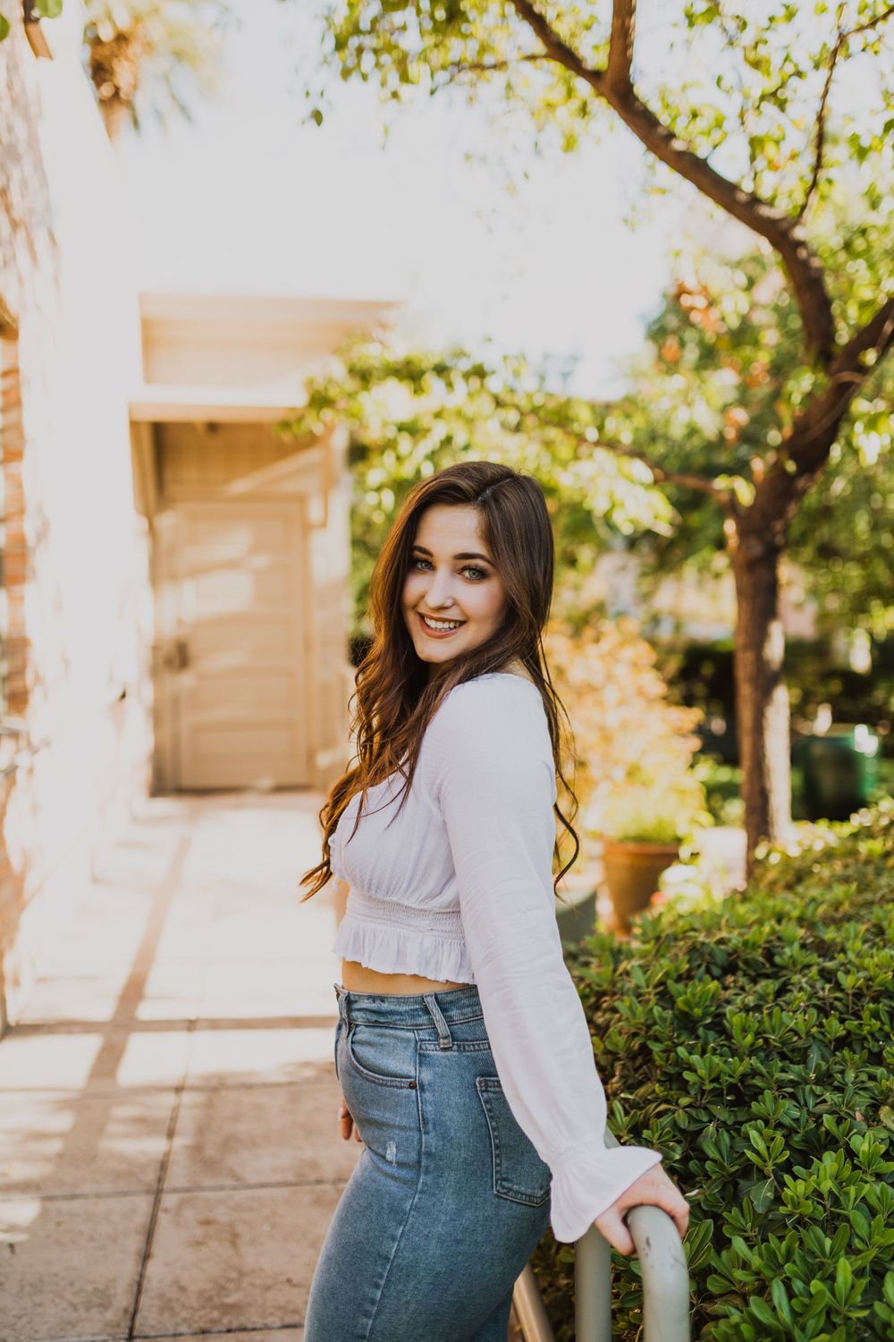 Casual outdoor photoshoot with white blouse and jeans against garden greenery.