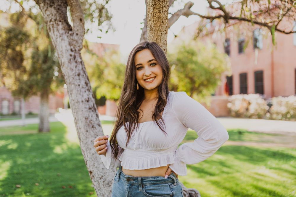 A person in a white ruffle top and jeans poses playfully in a sunny park setting with trees in the background.