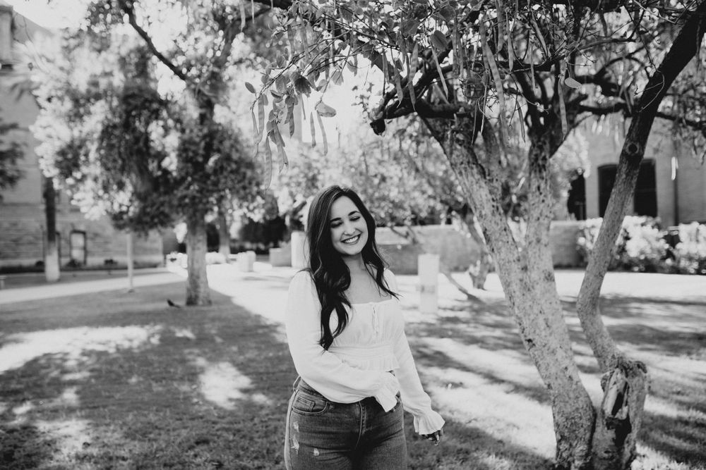 Black and white portrait of someone posing beneath tree branches in a relaxed outdoor setting.
