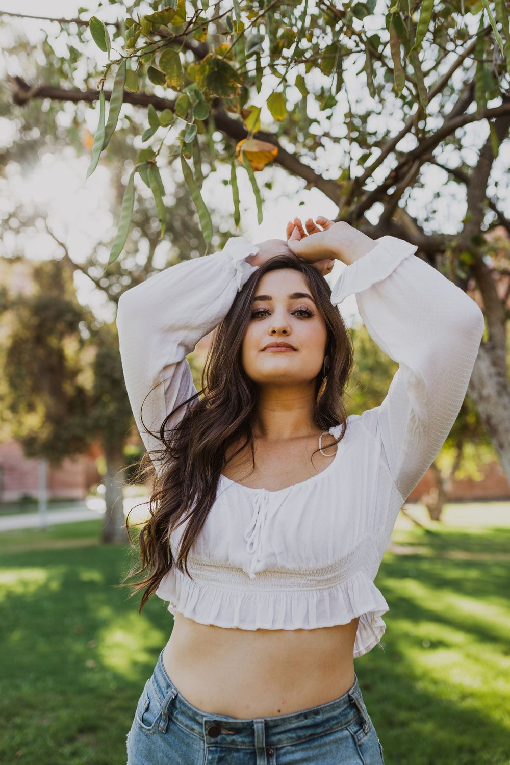 Lifestyle photography featuring someone in a cropped white top posing beneath trees in a park.