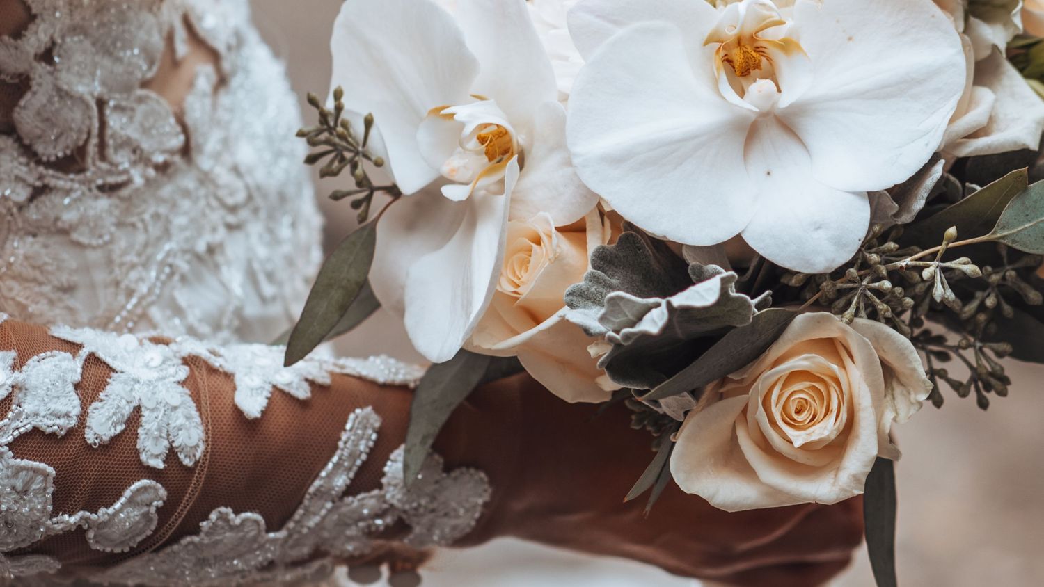 Bride's hand holding a white orchid, cream rose bouquet. Elegant lace dress detail. Wedding flowers in the Rockies.