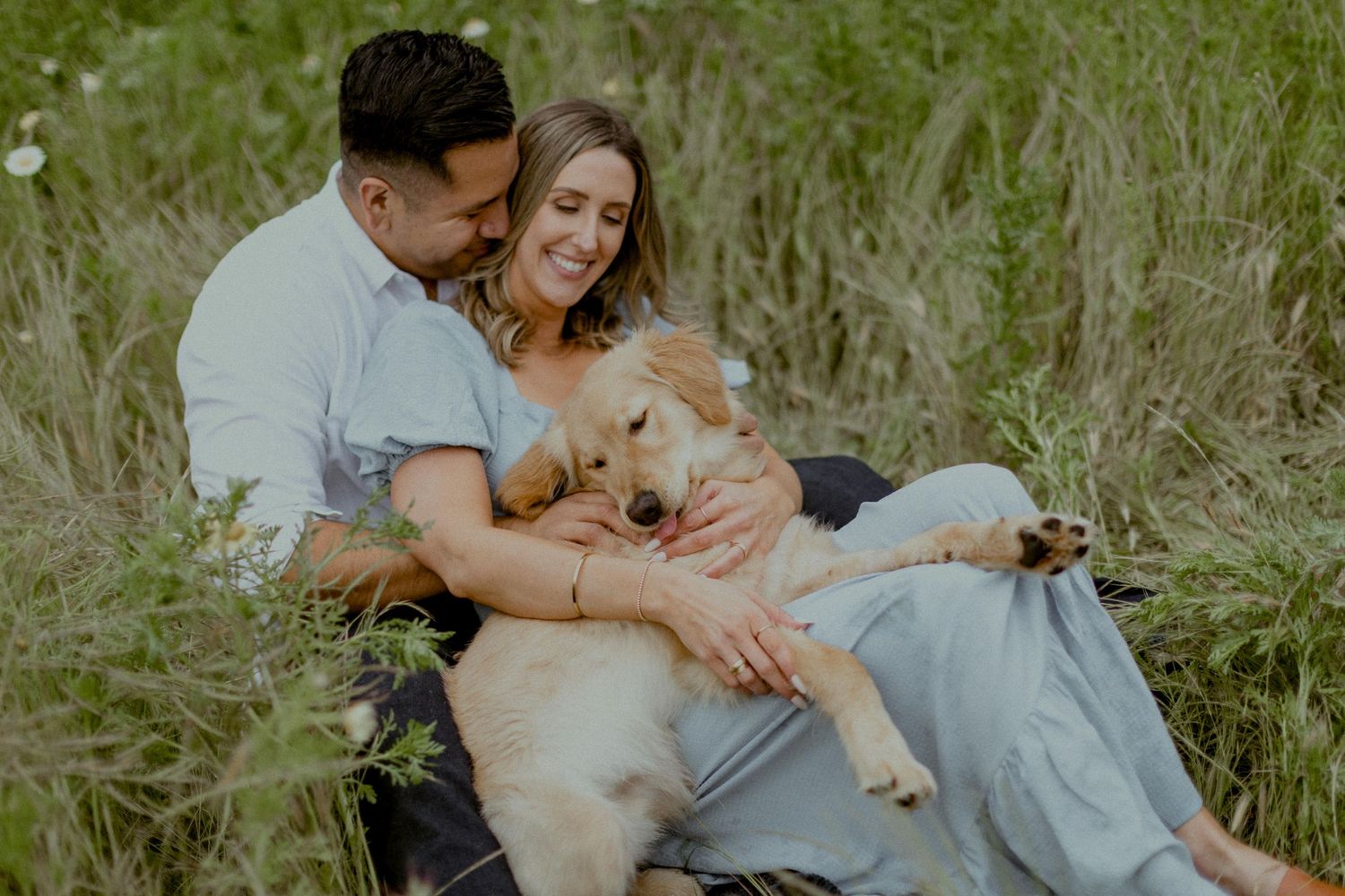 A happy couple cuddles with their golden retriever puppy in a grassy meadow.