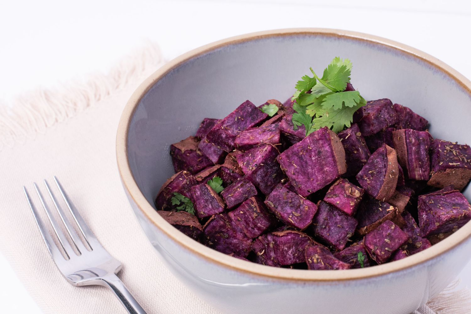 Roasted purple beet cubes with herbs in a grey bowl on white background with fork.