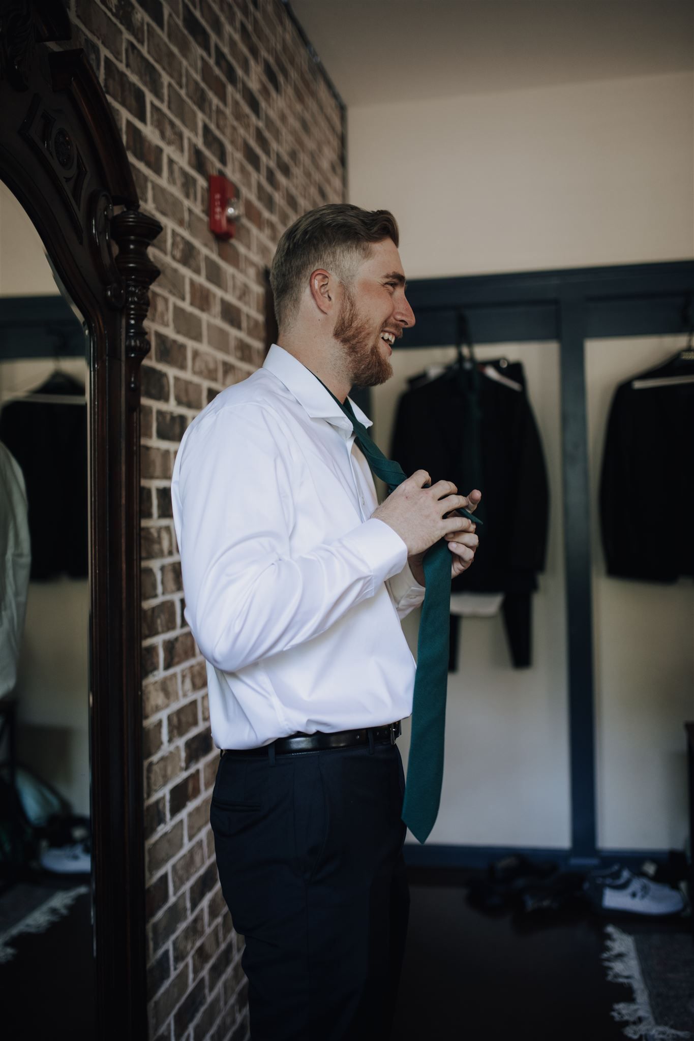 Groom in white shirt and bow tie smiles while getting ready in a brick-walled room.