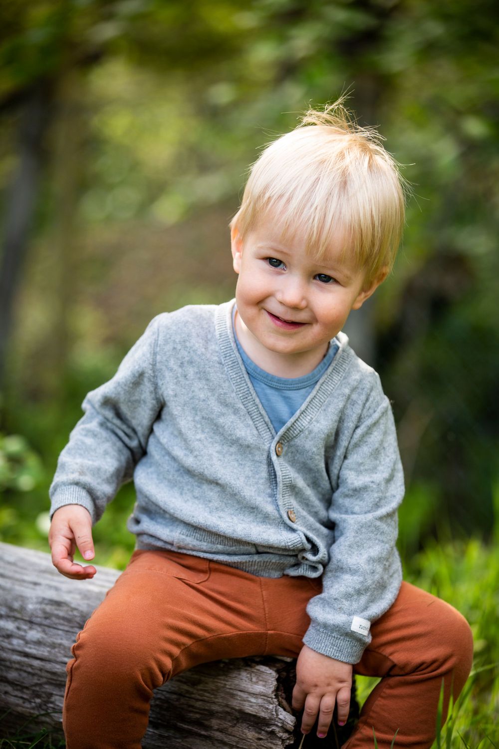 Child in gray sweater sitting on log with bright smile in outdoor setting.