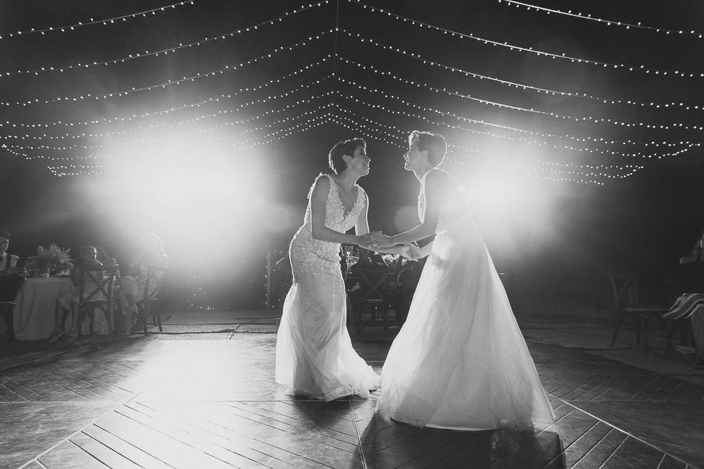 Two figures in white gowns share an elegant dance on a dimly lit wooden floor under twinkling string lights.