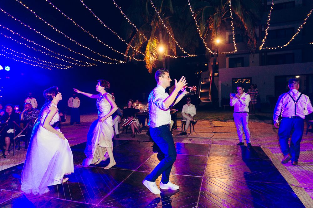 Wedding party dancing energetically on illuminated dance floor under strings of blue lights.