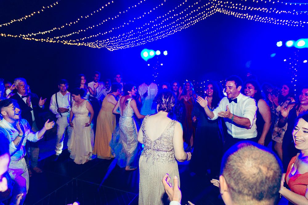 Crowded wedding reception dance floor illuminated by blue lights and overhead string lighting.