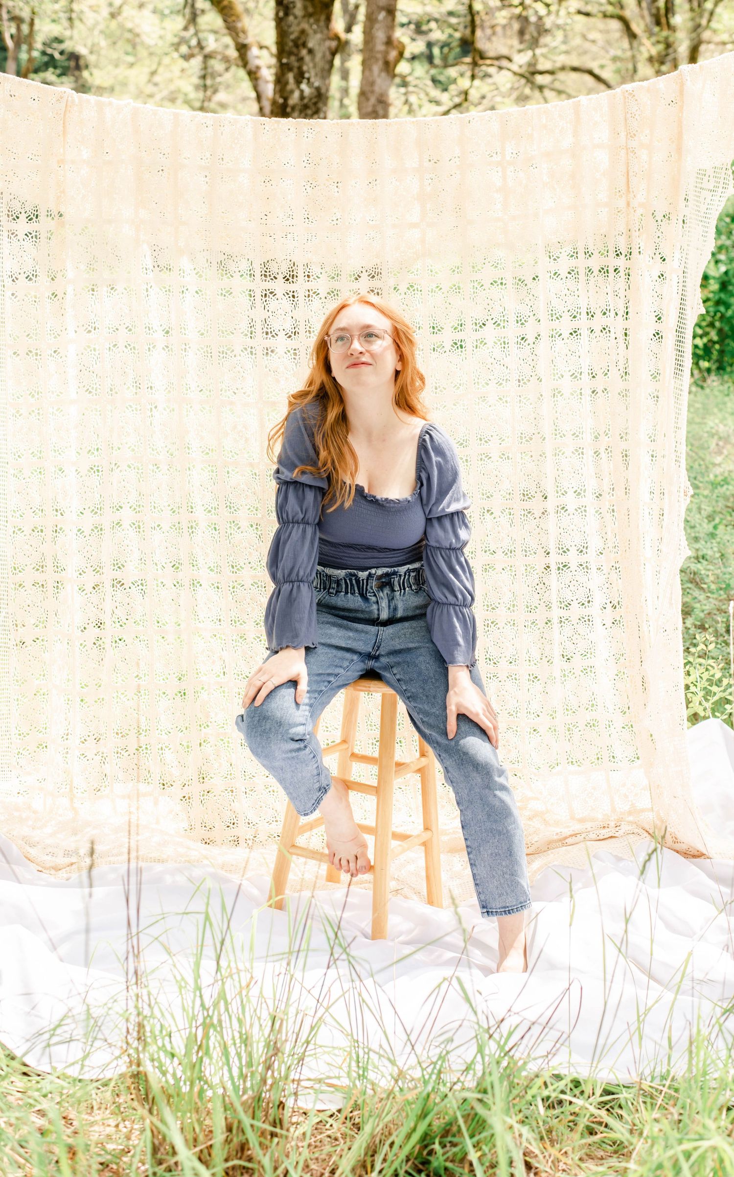 Person in blue dress sits on wooden stool against lace curtain backdrop outdoors.
