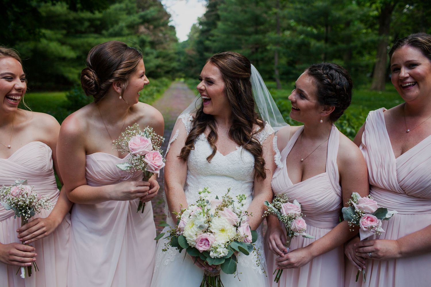 Bridesmaids in blush pink dresses share joyful moments together outdoors with delicate floral bouquets.