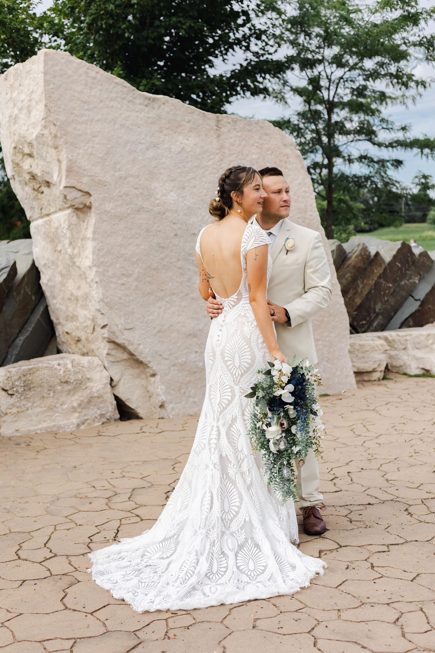 Bride and groom in elegant wedding attire posing against large stone backdrop with cascading bouquet.