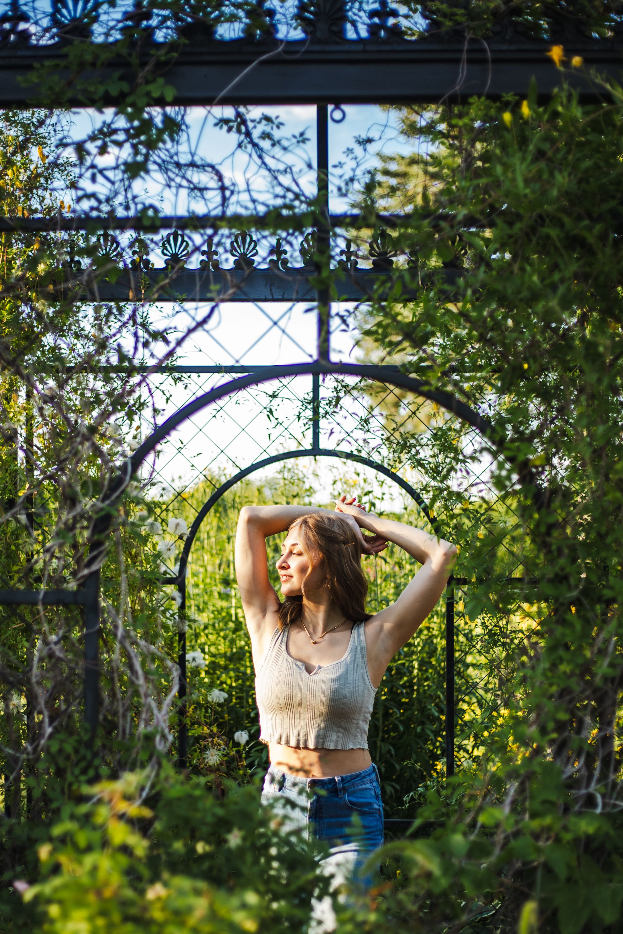 Someone poses under an ornate garden archway with sunlight filtering through trees.