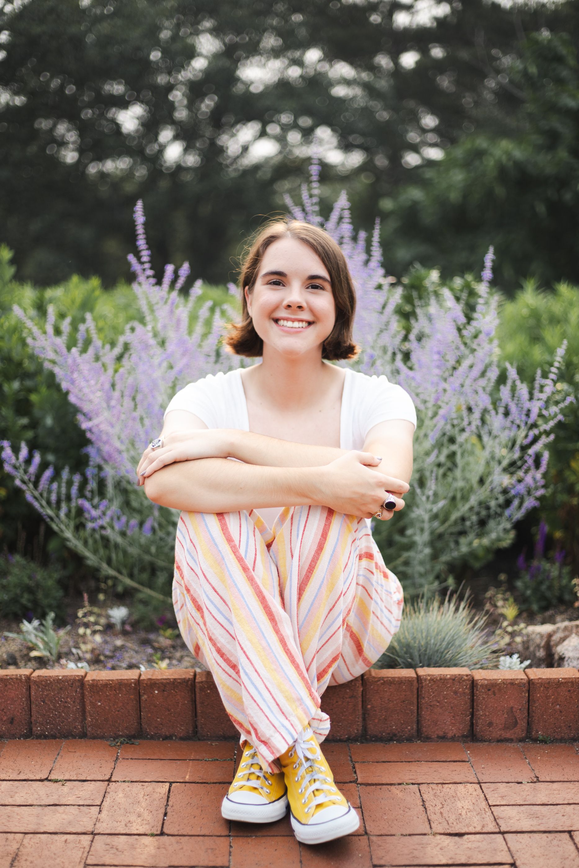 Someone sits on brick ledge surrounded by purple flowers wearing striped pants and yellow shoes.