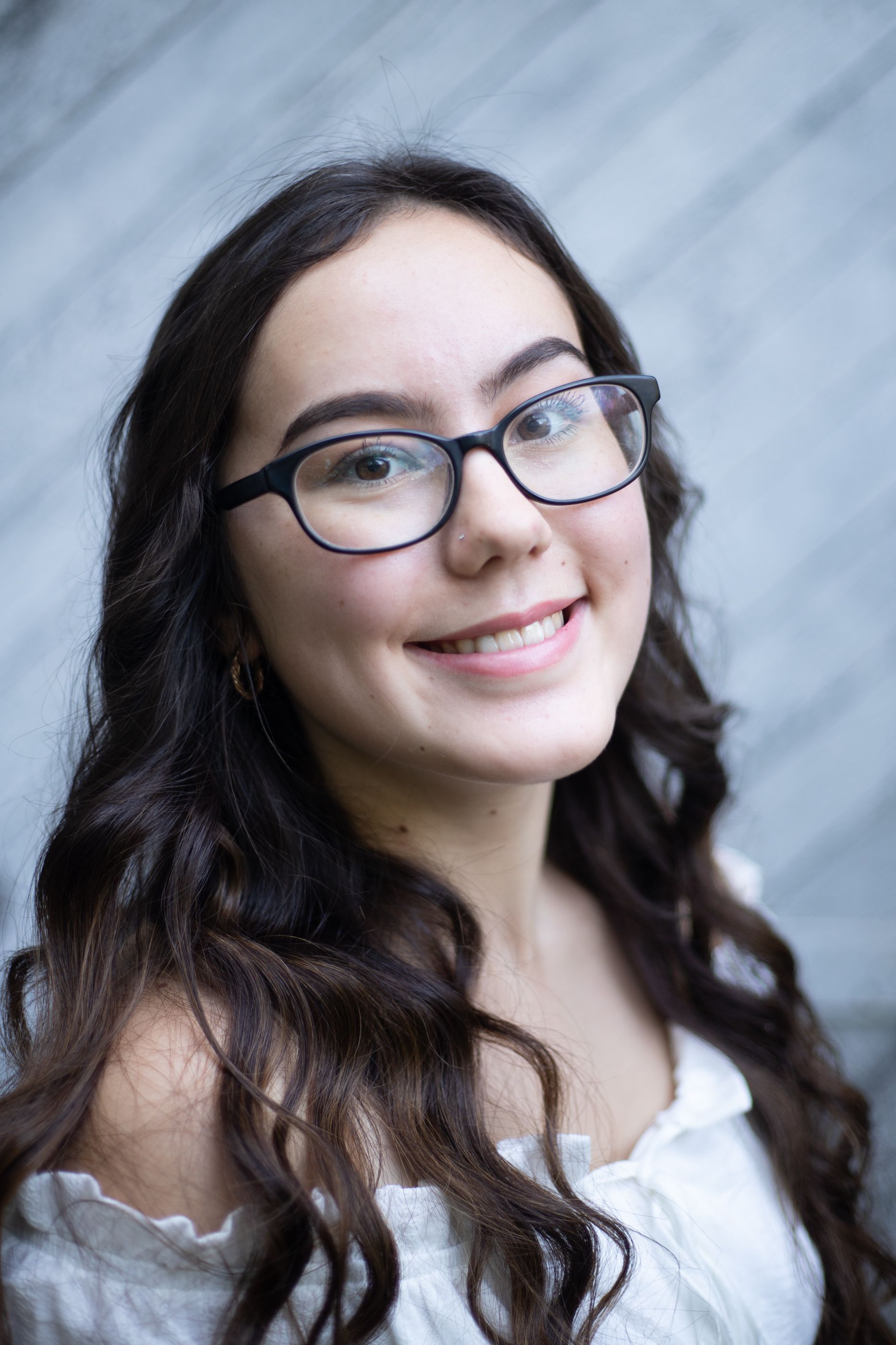 A close up portrait against a grey background features glasses and wavy dark hair.