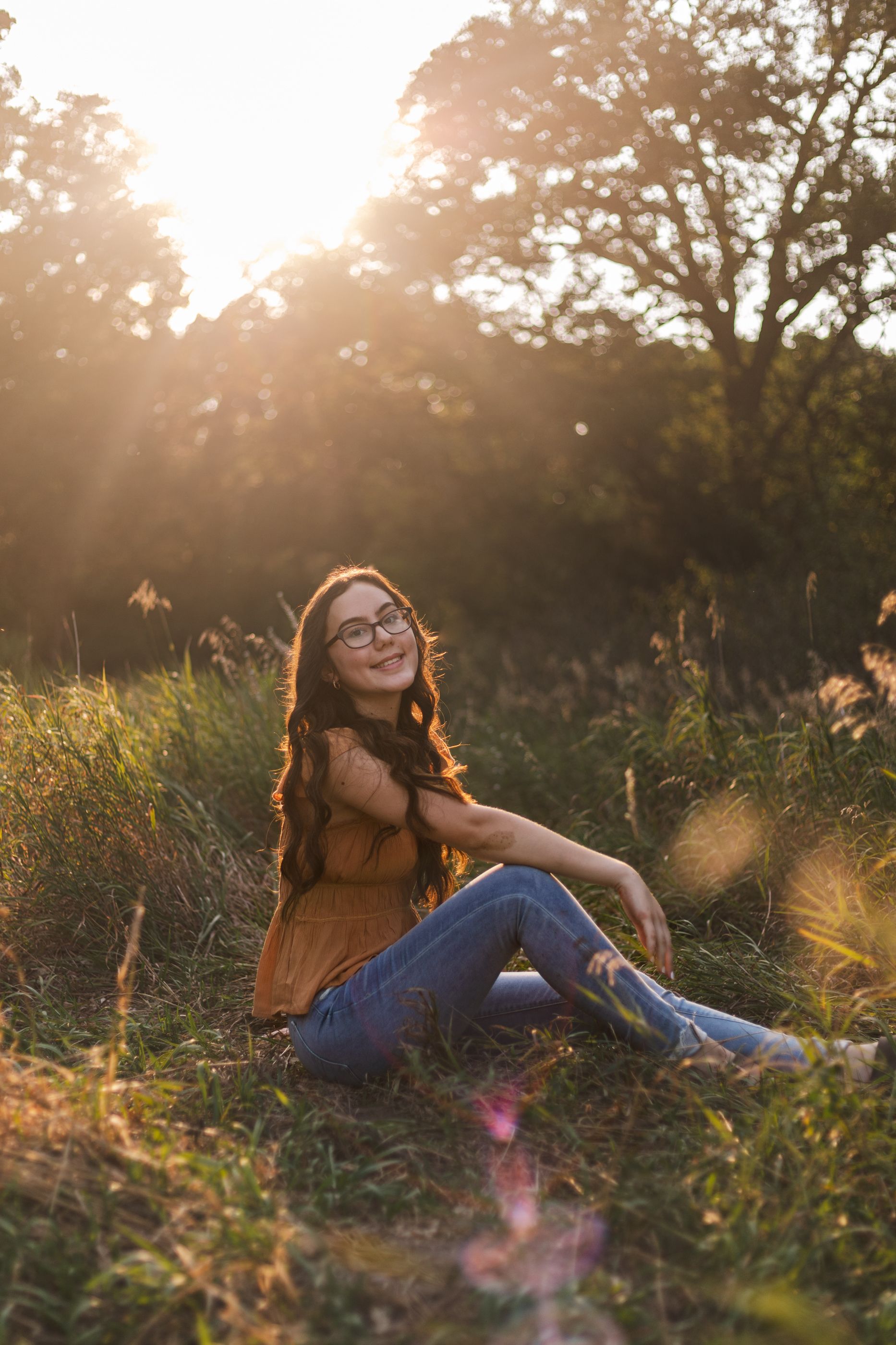 A candid sunset portrait in a field shows someone in a brown top and jeans relaxing in golden hour lighting.