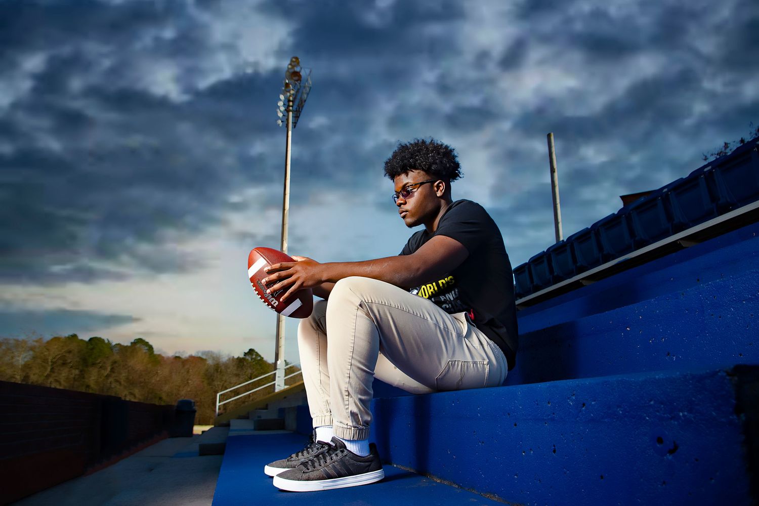 Athletic figure sits contemplatively on blue bleachers against dramatic stormy sky at dusk.