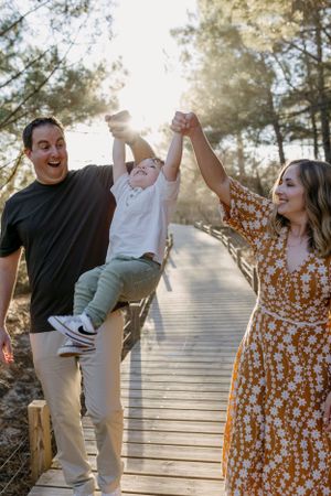 Playful family moment on wooden boardwalk surrounded by sunlit pine trees.