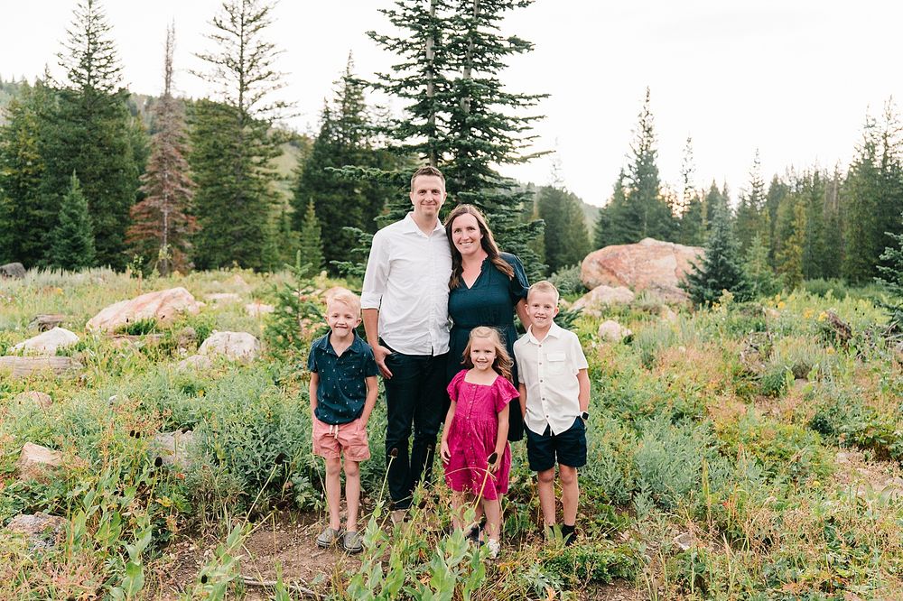 A family standing together in a mountain meadow with evergreen trees and summer wildflowers.