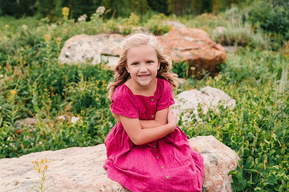 Child in bright pink dress sitting on rocky outcrop surrounded by mountain wildflowers.