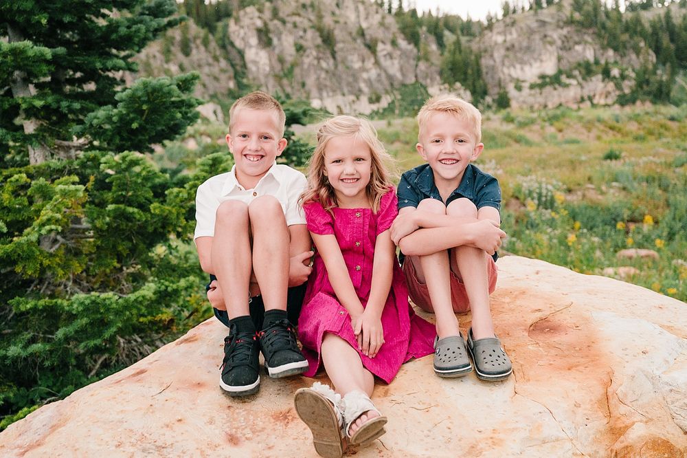 Three children sitting together on a large red rock with mountain scenery in the background.