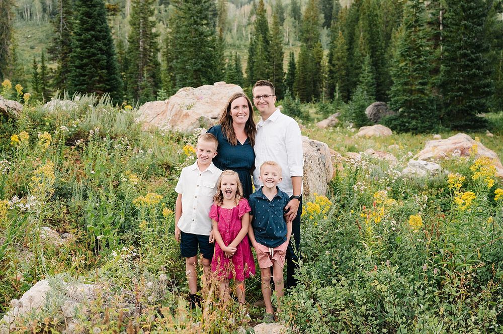 Smiling group poses together in mountain meadow with evergreen forest and yellow wildflowers.