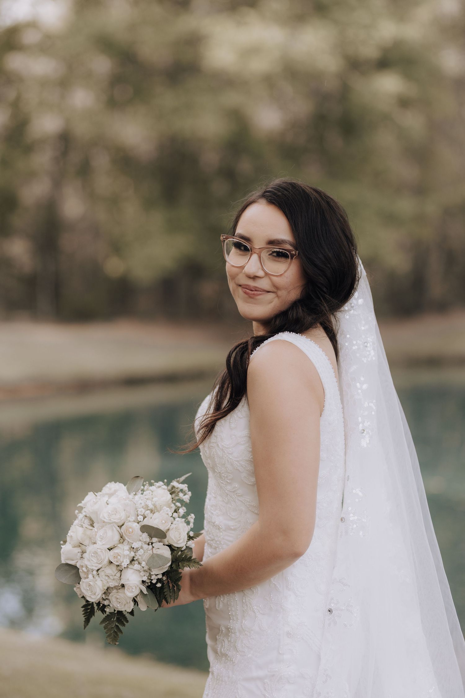Bride with glasses holds white bouquet while posing near water with long veil flowing behind her.