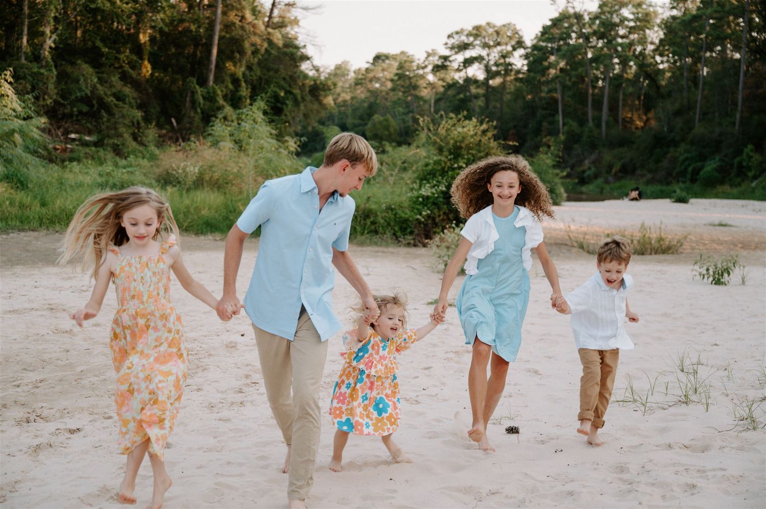 Family holding hands while running and playing on sandy beach path during sunset.