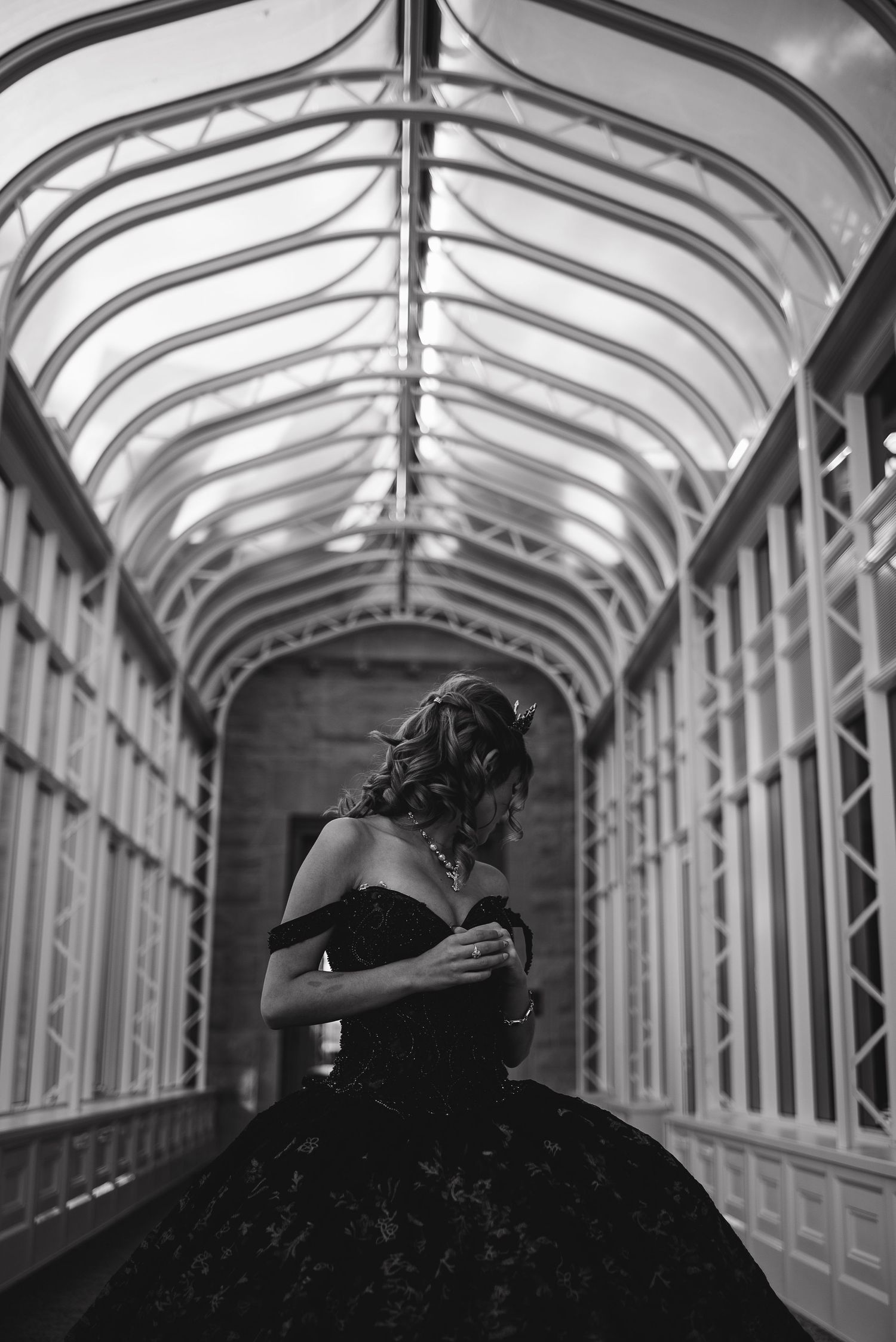 Dramatic black and white portrait of figure in ballgown under arched architectural ceiling.