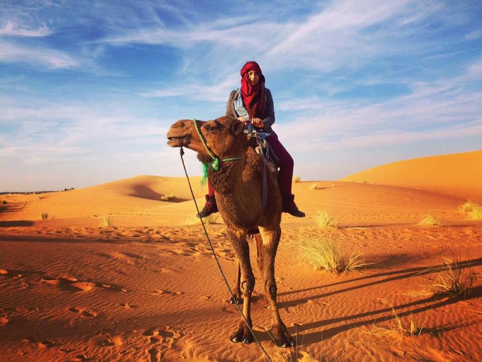 A woman on a camel in Sahara Desert Erg Chebbi Merzouga, Morocco. Sand dunes and sky behind her. She is wearing a hijab